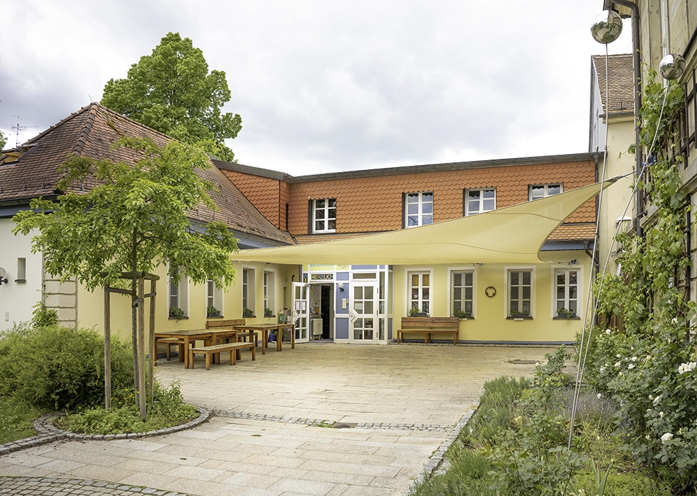 Innenhof des Integrativen Montessori Kindergartens Bayreuth mit Eingangsterrasse, Sonnensegel und Bäumen