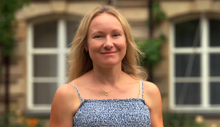 Porträt von Christina Weidl, Kinderpflegerin im Integrativen Montessori Kindergarten Bayreuth, mit langen blonden Haaren und kariertem Overshirt vor grünem Gartenhintergrund