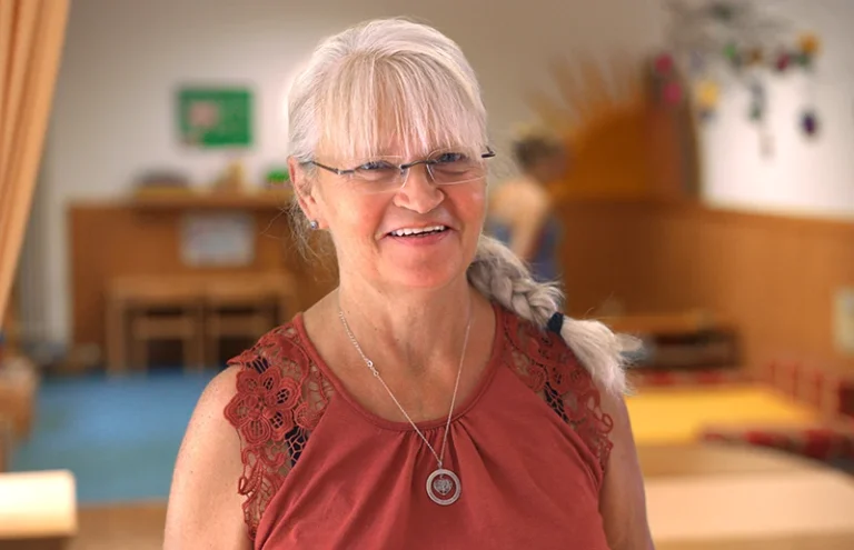Porträt von Heide Hientz, Köchin und Raumpflegerin im Integrativen Montessori Kindergarten Bayreuth, mit blondem Haar, Brille und Perlenkette vor blauem Hintergrund